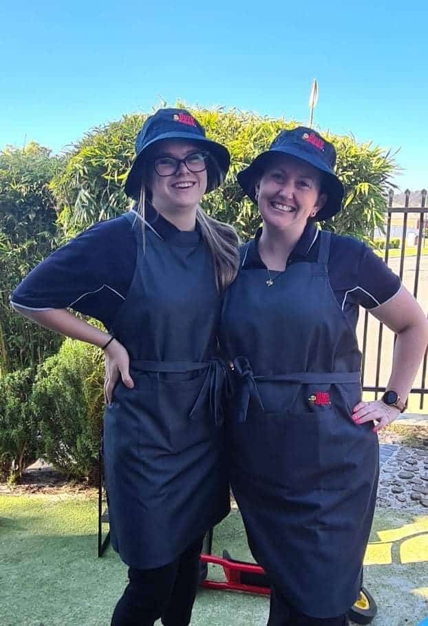 Two people in gray aprons and bucket hats pose happily outside in front of a hedge, capturing the joyful spirit of early learning.