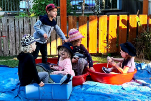 Five children from the day care are playing outdoors, some in plastic tubs while others stand nearby. They are under a wooden structure with a wooden fence in the background.