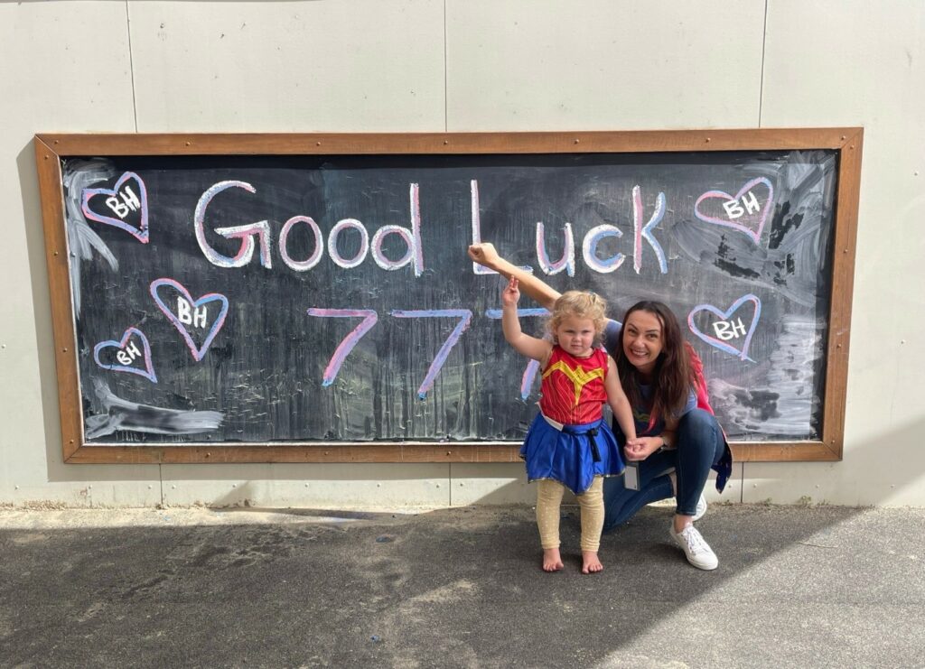 Two people stand in front of a chalkboard with "Good Luck 77!" surrounded by heart drawings. The child, dressed in a superhero costume, beams with excitement as the adult from their kindergarten points at the encouraging message.