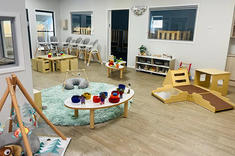 A modern, well-lit childcare room with various play areas and educational toys, including a play mat, a small slide, a table with toy food, and a row of high chairs by the window. Perfect for preschool or kindergarten activities.