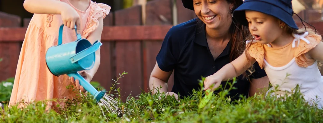 Buzzworthy Feature_Veggie & Herb Garden Two young girls, wearing hats and aprons, water plants in a garden bed while a woman kneels beside them, smiling and assisting. It's a delightful moment of early learning and childcare in nature's classroom.
