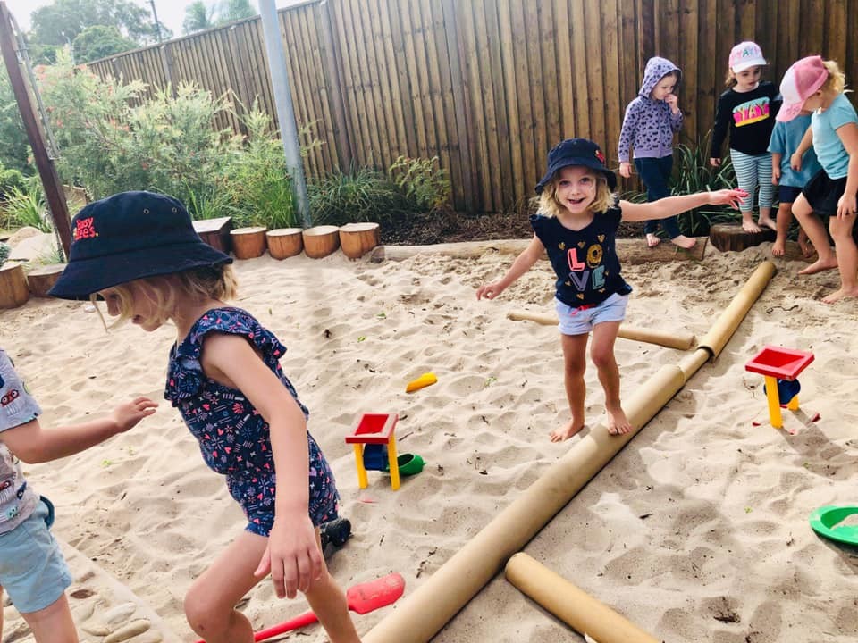 Photograph of children balancing on a log in the sandpit. The children are smiling and enjoying the play, taking turns to cross the log. The location is Busy Bees at Burpengary East.