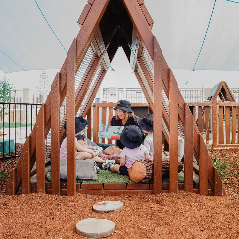 Busy Bees Early Childhood Educator reading a book to children inside a timber a-frame tent located in the outdoor playground of Busy Bees at Quinns Beach