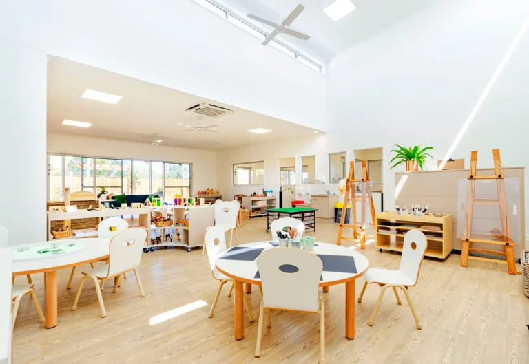 A brightly lit kindergarten classroom at Busy Bees Burpengary East with round tables, chairs, easels, and various educational materials arranged neatly.