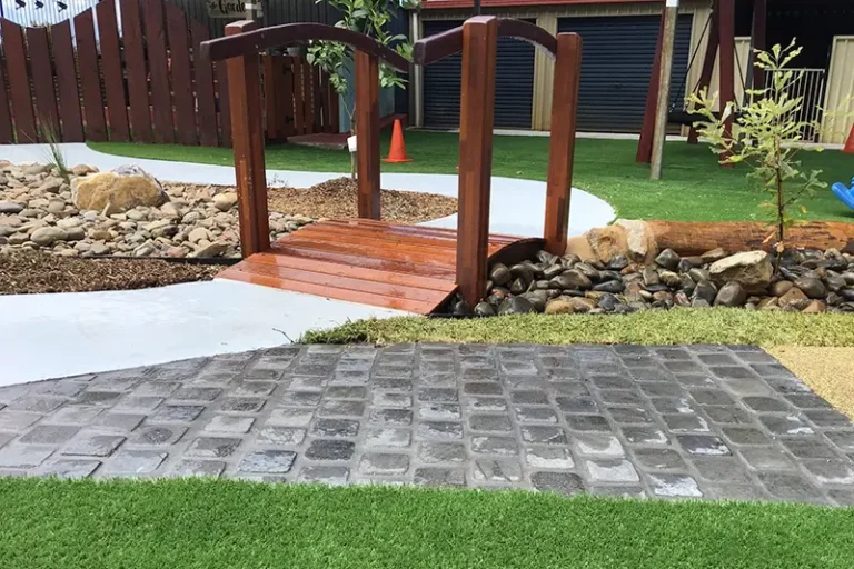 A small wooden footbridge over a decorative dry stream bed, surrounded by a pathway, rocks, and artificial grass in a landscaped yard provides a charming outdoor space perfect for preschool or childcare activities.