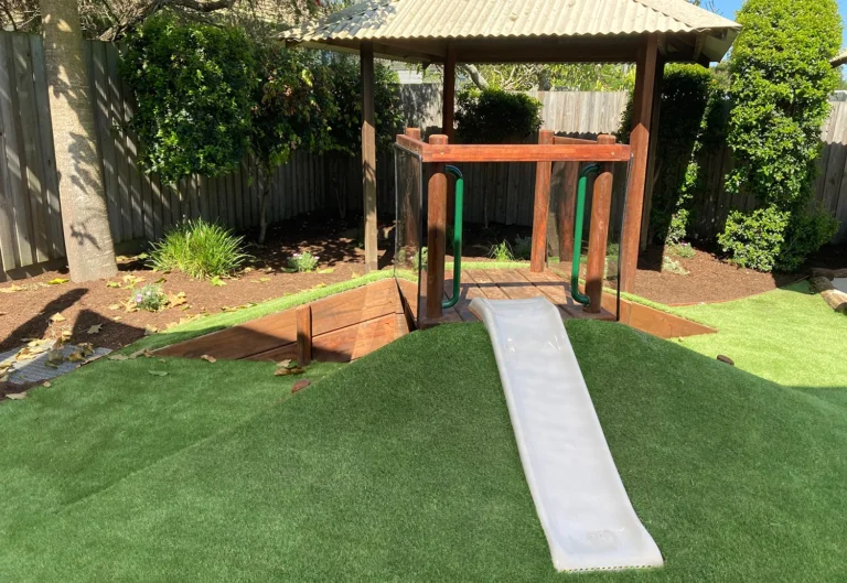 Nursery playground at a day care in Beenleigh featuring a wooden play structure with a small white slide siting on a grassy mound.