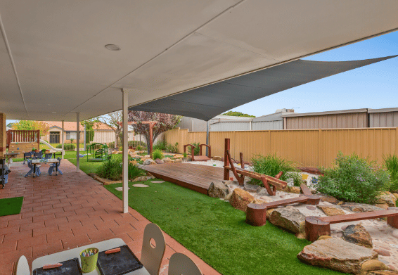 Outdoor nature play area with artificial grass, rocks, garden beds and timber balance elements.