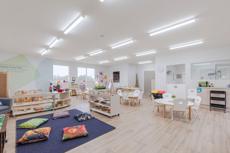 A bright, modern preschool classroom with several activity stations, small tables and chairs, pillows on a rug, and shelves filled with educational materials. The walls are mostly white with some colorful decor.