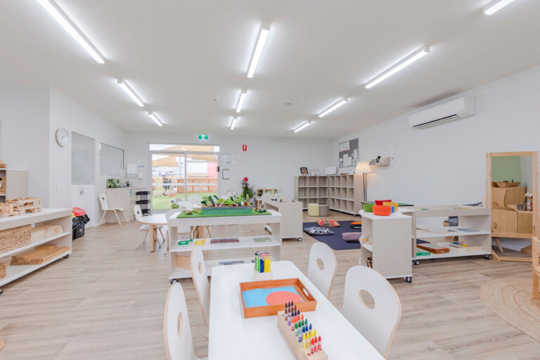 A brightly lit, neatly arranged kindergarten classroom with child-sized furniture and various educational materials on tables and shelves. Large windows and a door open to an outdoor space, creating an ideal environment for early learning.