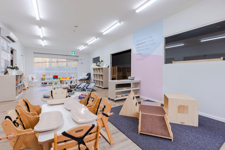 A bright, well-lit daycare room with wooden furniture, high chairs, toys, and a play area. The walls are decorated with posters and quotes promoting early learning to create a nurturing environment for childcare.