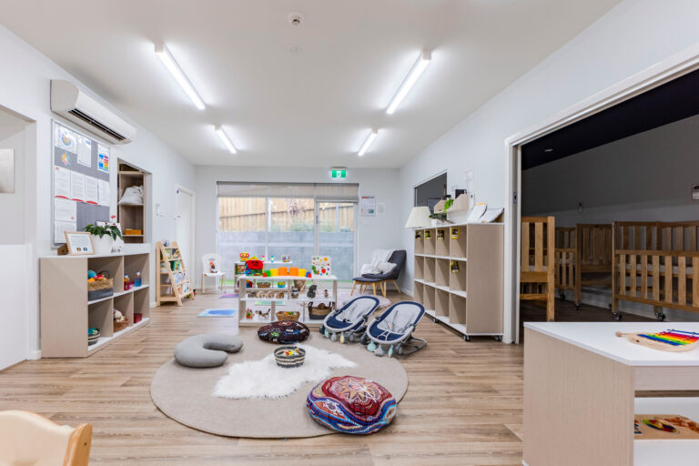 A brightly lit, organized preschool room with various toys, books, activity stations, and seating areas. Cribs are visible through an open door to an adjoining childcare room.