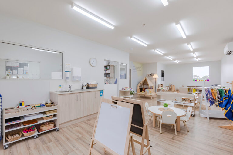 A brightly lit preschool classroom with wooden floors, tables, chairs, a play kitchen, and storage shelves. There is a sink, a clock on the wall, and a window providing natural light. The space is neatly organized to offer an ideal environment for childcare activities.