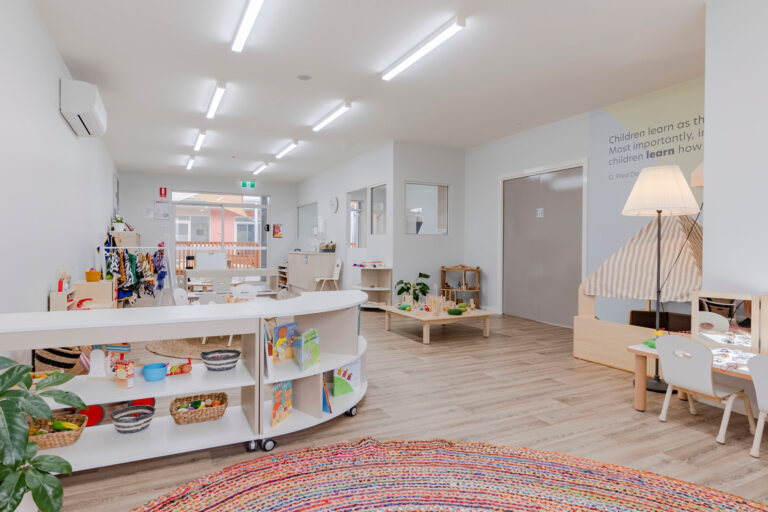 A well-lit, neatly organized preschool classroom with various learning stations, bookshelves, a reading nook, and a colorful rug in the center. Educational materials are arranged on shelves, and there are tables for activities.