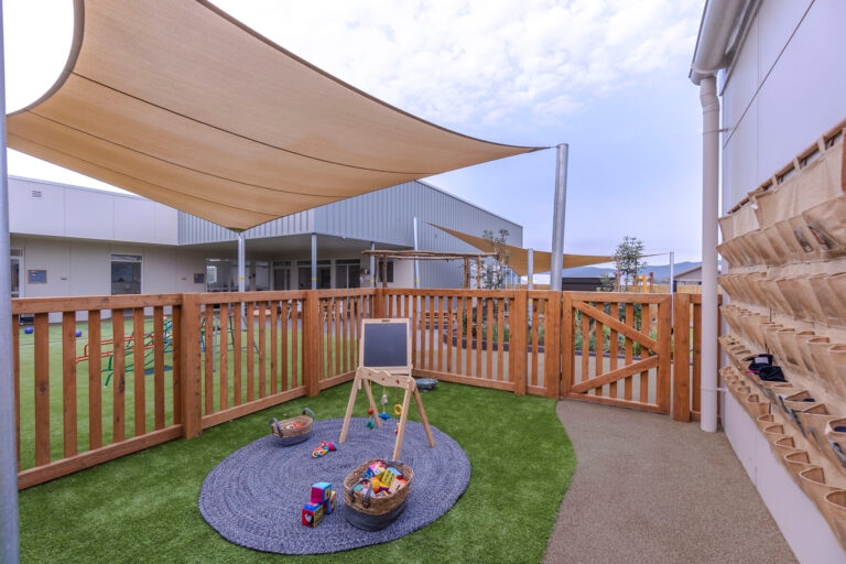A fenced outdoor play area at the preschool features a rug and toys on artificial grass. An easel stands in the middle, while a structure overhead provides shade. The background reveals buildings and a cloudy sky, creating an inviting space for childcare activities.