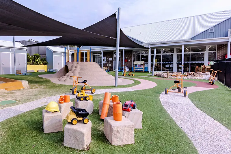 A playground with various toys, including tricycles and construction vehicles, scattered around. Shade sails cover parts of the area, and a mound structure with a slide is visible in the background.