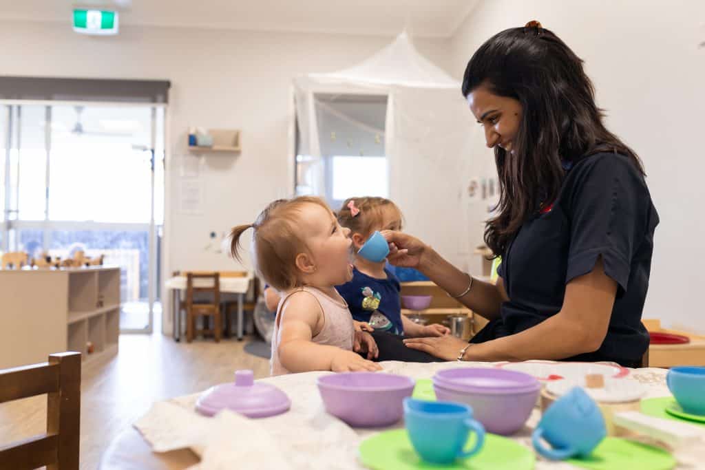 In a cozy preschool room set up with toy kitchen items, a caregiver helps a young child drink from a small cup, while another child sits in the background.