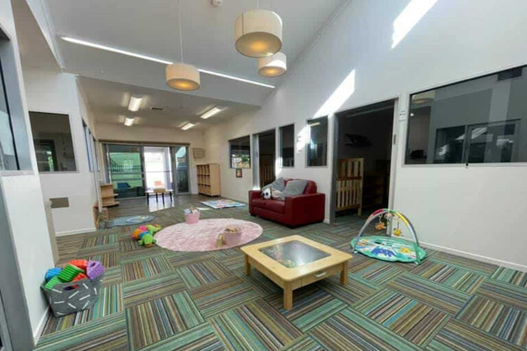A brightly lit daycare room with toys including a playmat, foam blocks, and an activity gym. A red couch and small tables are placed around the room with large windows on the walls, creating a perfect environment for early learning.