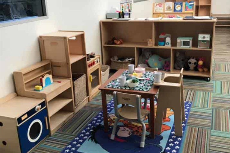 A tidy children's playroom with wooden shelves and a small table. The table is set with toy dishes, and the floor has a colorful rug. Shelves hold various toys and books, perfect for early learning in a preschool or childcare setting.