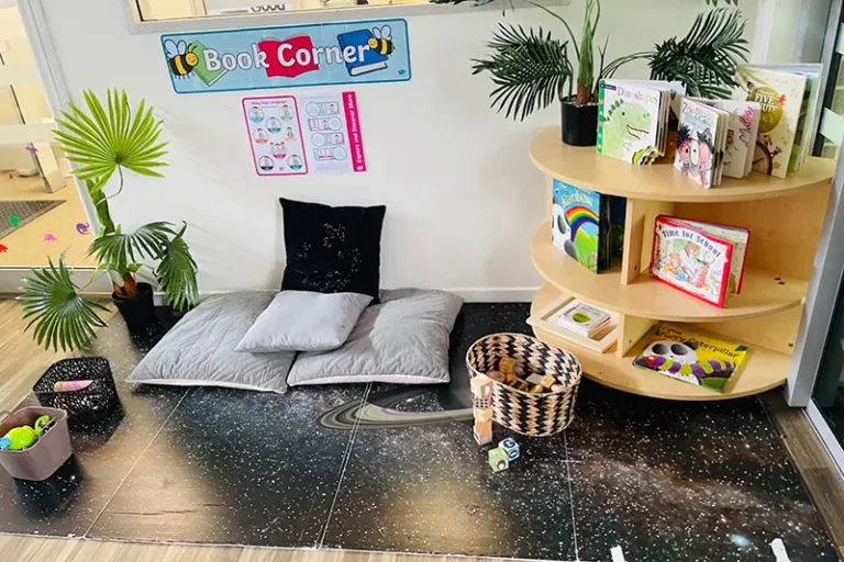 A cozy reading nook with cushions, a round shelf filled with books, and potted plants. A sign above reads "Book Corner." Baskets with toys are placed on the space-themed mat, perfect for day care or early learning environments.