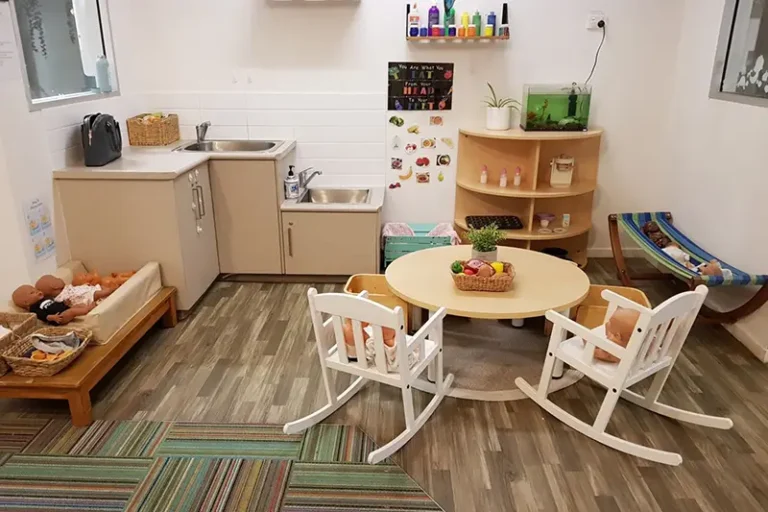 Image of a small, tidy daycare playroom with a round table, two white rocking chairs, a sink, shelves with toys, a play kitchen, and a hammock. The floor has a mix of tile and carpet.