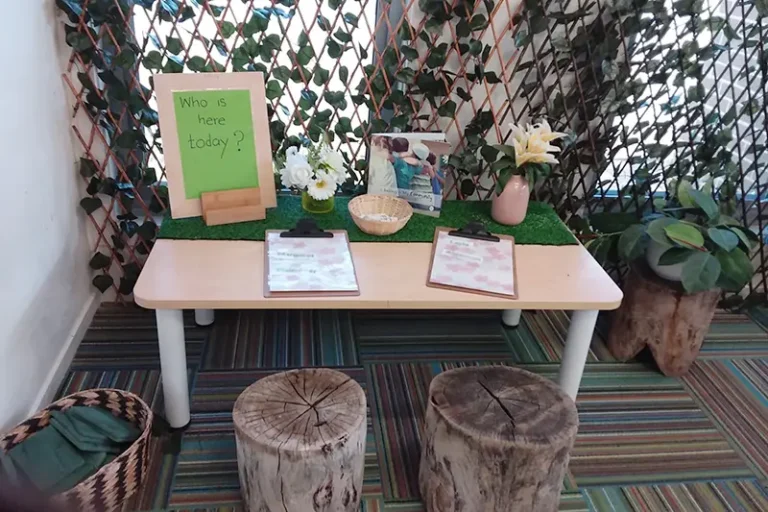 A small table with clipboards, a basket, a vase of flowers, and a framed photo creates a welcoming atmosphere in the kindergarten. A green board asking "Who is here today?" adds to the early learning environment, while wooden stools and green plants enhance the cozy childcare space.