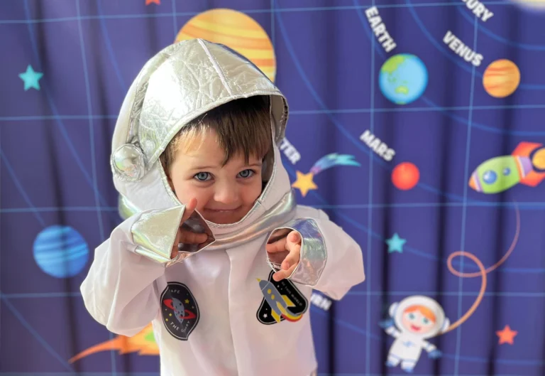 A child from Busy Bees Chinchilla in a white astronaut costume with a silver helmet poses in front of a space-themed backdrop featuring planets and stars.