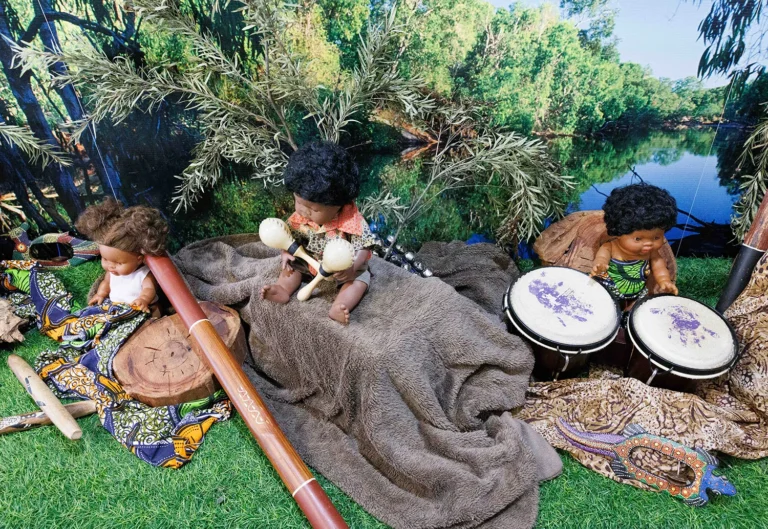 Three dolls sit on a grassy surface surrounded by musical instruments, including a didgeridoo, maracas, and bongos, with a nature backdrop of trees and a pond.