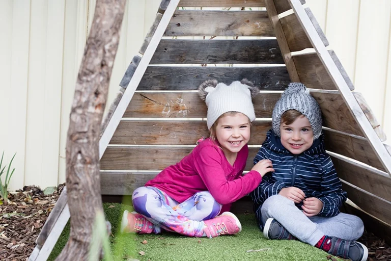 Two young children sit together inside a small wooden structure. Both wear beanies and smile at the camera, with the child on the left in pink and the child on the right in blue.