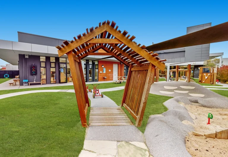 Wooden structure in a modern playground with green grass, sandpit, tricycles, and a building in the background under a clear blue sky.