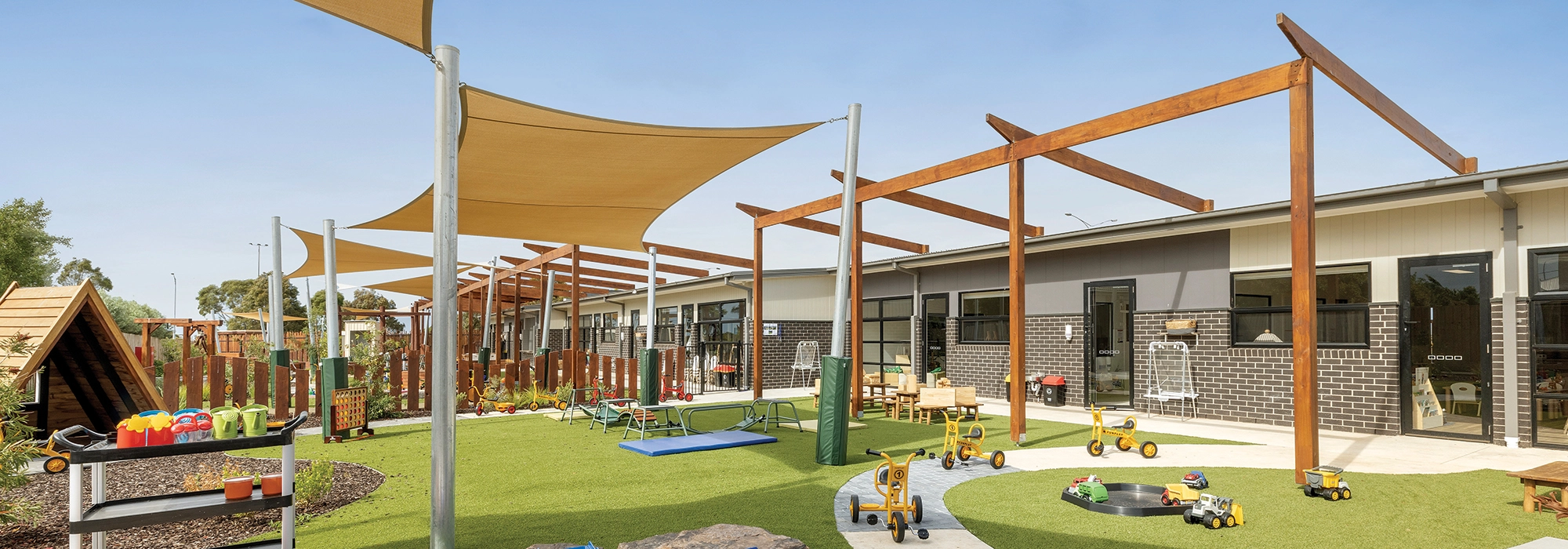 A well-equipped outdoor play area at a daycare center, featuring shade canopies, tricycles, toys, a small playground, and various activity stations amidst a grass and concrete surface.