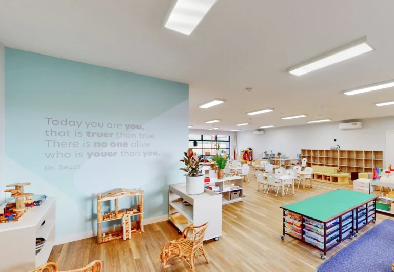 A brightly lit classroom with a Dr. Seuss quote on the wall, featuring tables, chairs, storage shelves, and various educational materials and toys.