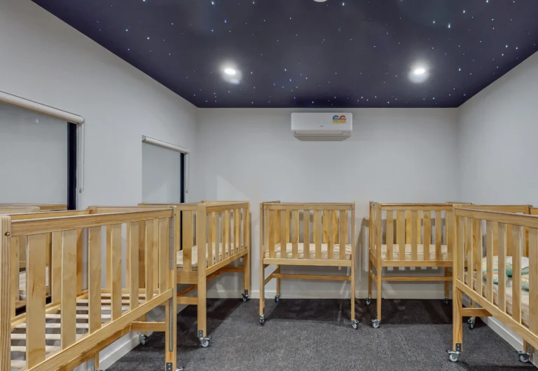A room with wooden cribs arranged in a row, featuring a dark ceiling with star-like lights.