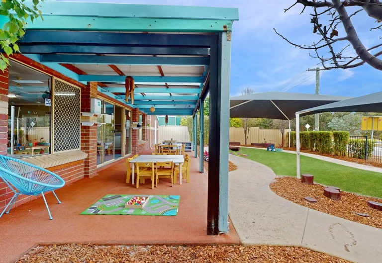 A covered outdoor area at Byford North daycare center featuring tables and chairs, a play mat with toys, and surrounding green space with pathways and a canopy.
