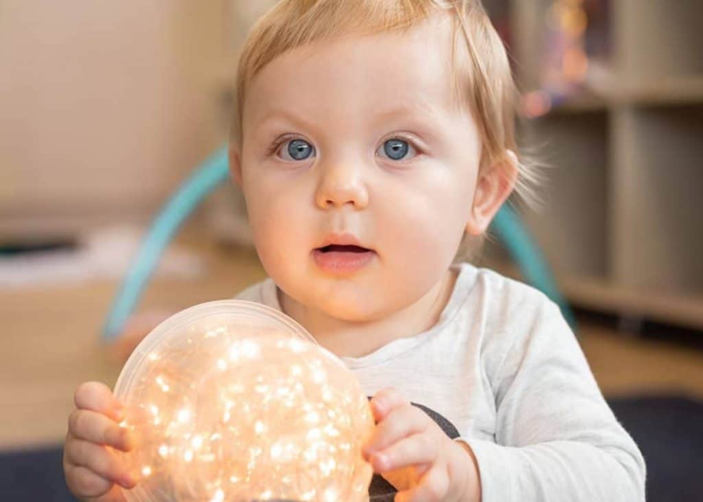 A baby with blue eyes holds a glowing ball of lights while looking ahead, in a vibrant kindergarten setting.