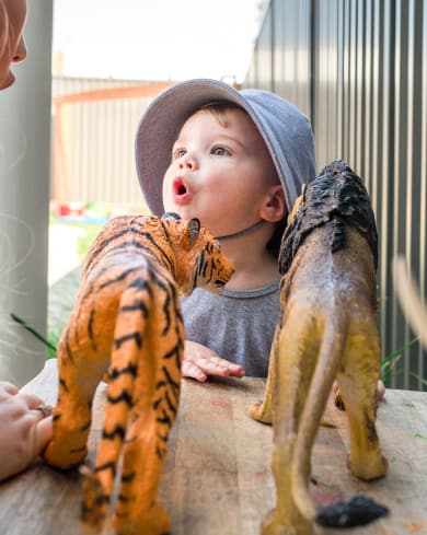 A young child in a blue hat interacts with two toy animal figures, a tiger and a lion, on a wooden surface outdoors at their preschool.