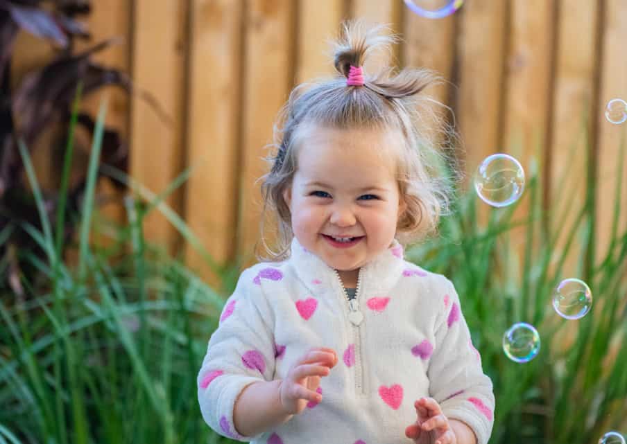 A young child with light hair in a topknot, wearing a white jacket with pink hearts, stands outdoors near green plants and wooden fencing at their preschool, smiling and looking at floating bubbles.