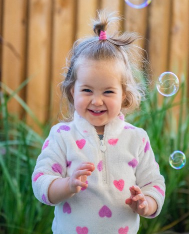 A toddler with a topknot hairstyle wearing a white sweater with pink hearts, smiling and reaching towards bubbles outdoors, embodies the joy of early learning and discovery.