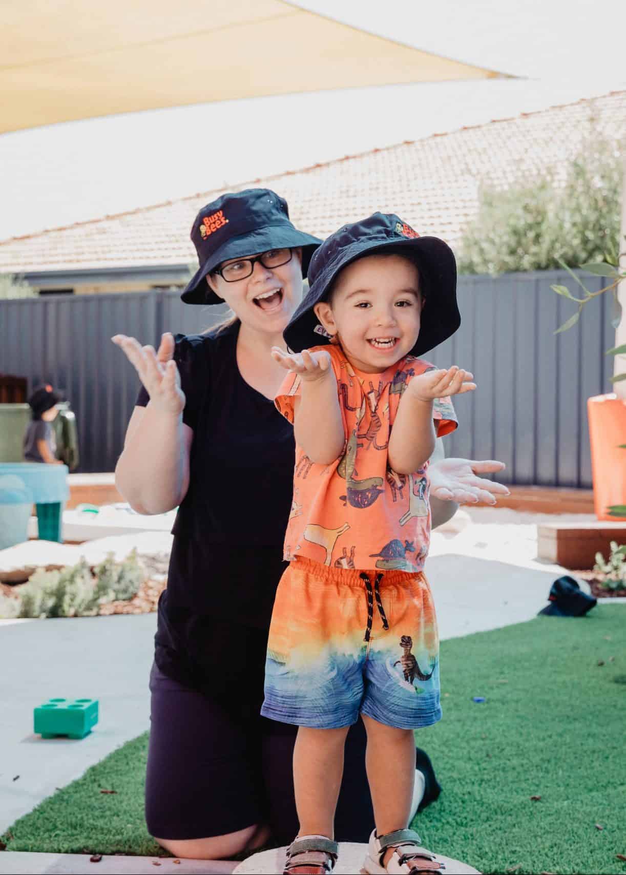 An adult and a young child wearing sun hats smile and gesture gleefully in an outdoor setting, with a preschool playground area in the background.
