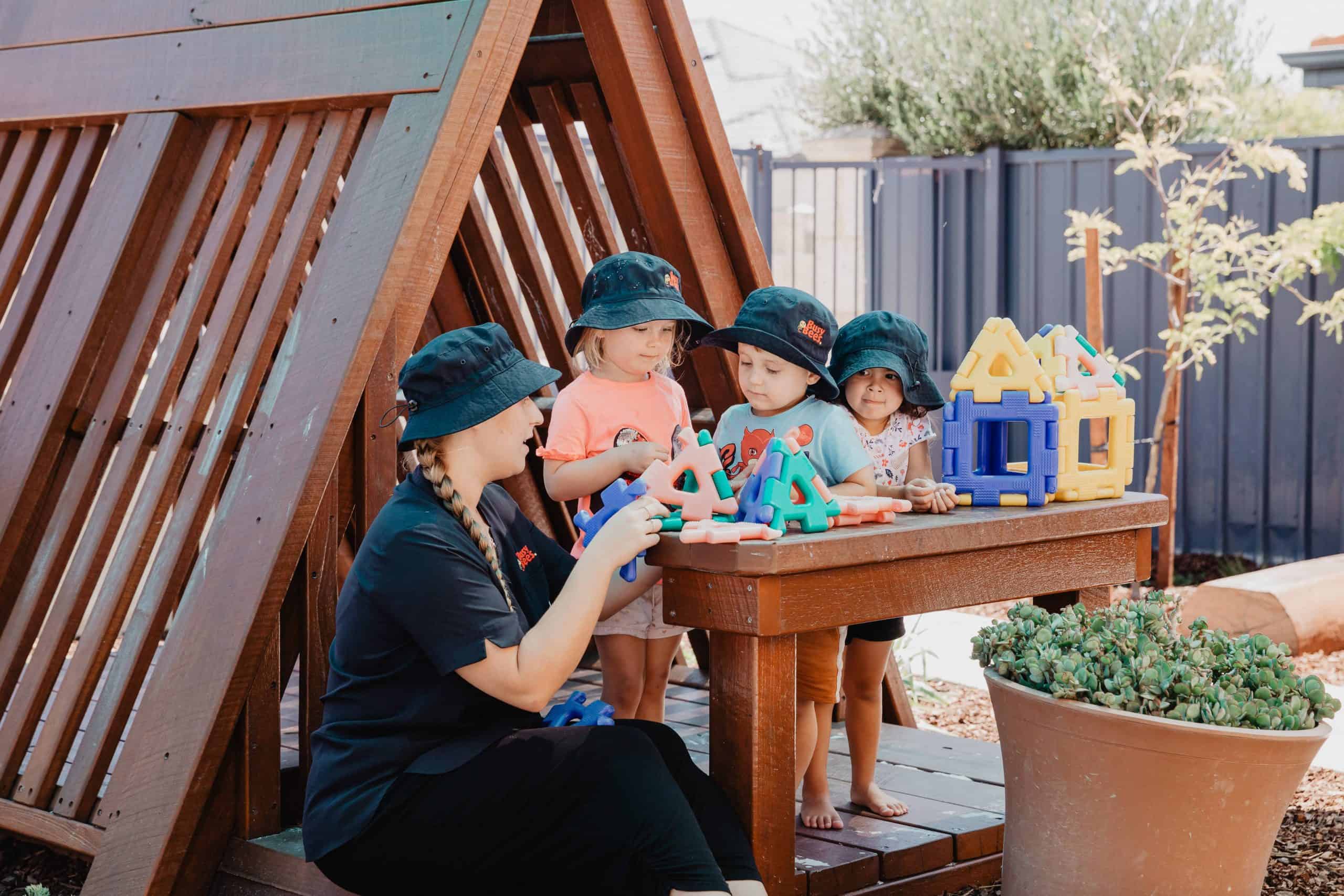 A preschool teacher and four children with hats play with colorful building toys at a wooden table outdoors by a triangular structure.