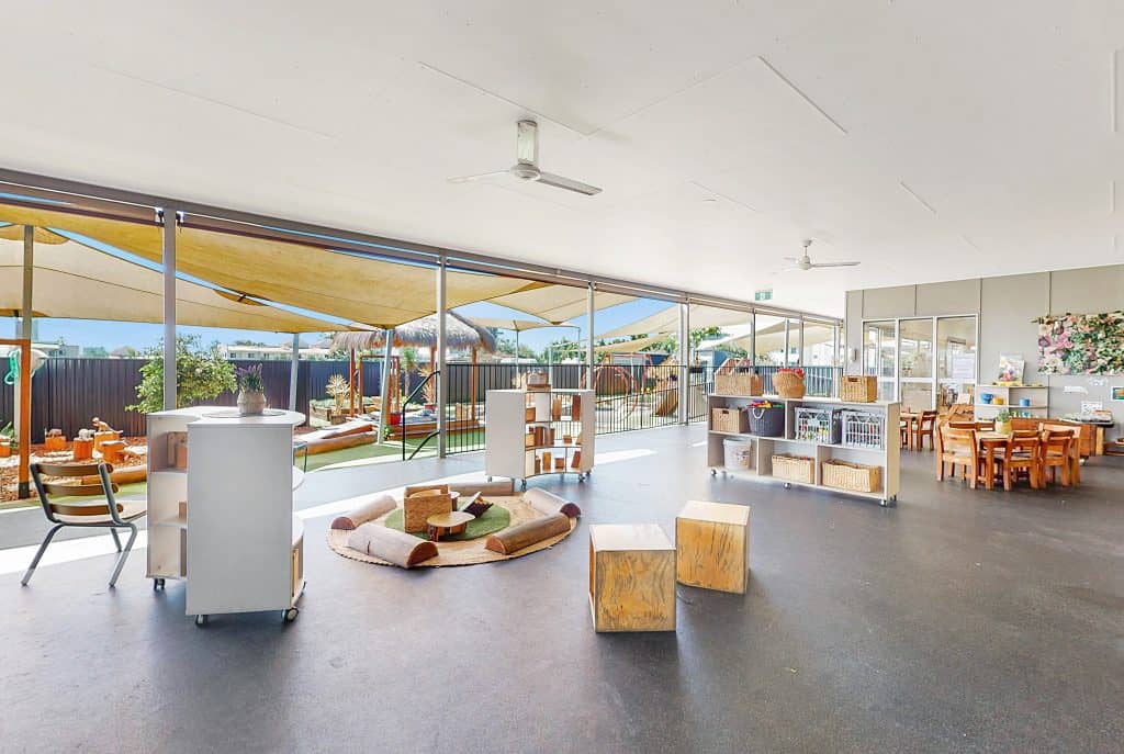 A spacious, well-lit classroom designed for early learning with large windows, wooden furniture, and various educational materials. An outdoor play area is visible through the windows, making it an ideal setting for kindergarten and childcare activities.