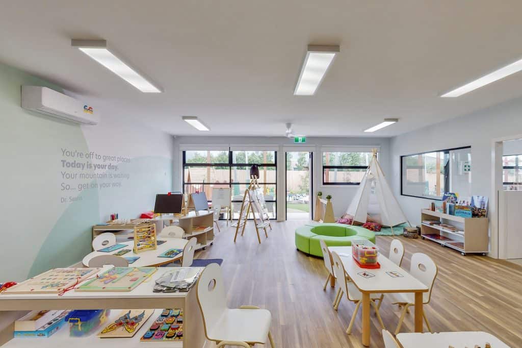 A bright, modern preschool classroom with wooden floors, child-sized furniture, bookshelves, a teepee, an art easel, and large windows. Various educational materials and decorations are neatly organized throughout. Perfect for fostering young minds in a nurturing childcare environment.