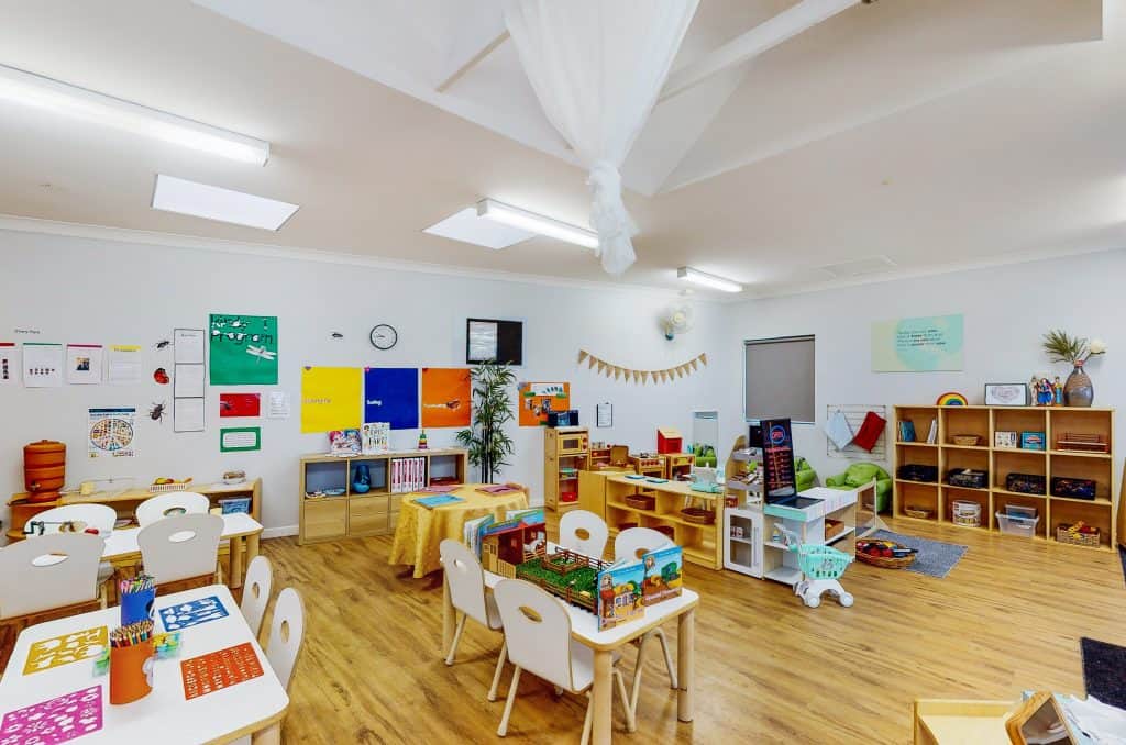 A brightly lit preschool classroom with tables, chairs, bookshelves filled with educational materials, colorful artwork on the walls, and a variety of toys and early learning stations.
