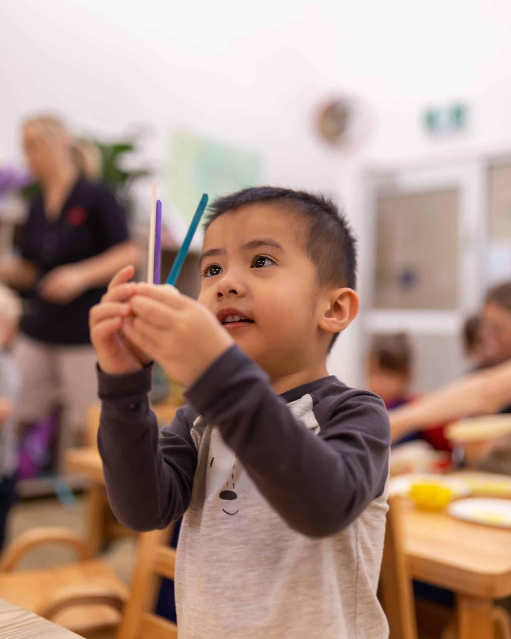A young child in a preschool classroom holds up two colored pencils and appears focused. Other children and an adult are visible in the background, creating a vibrant early learning environment.