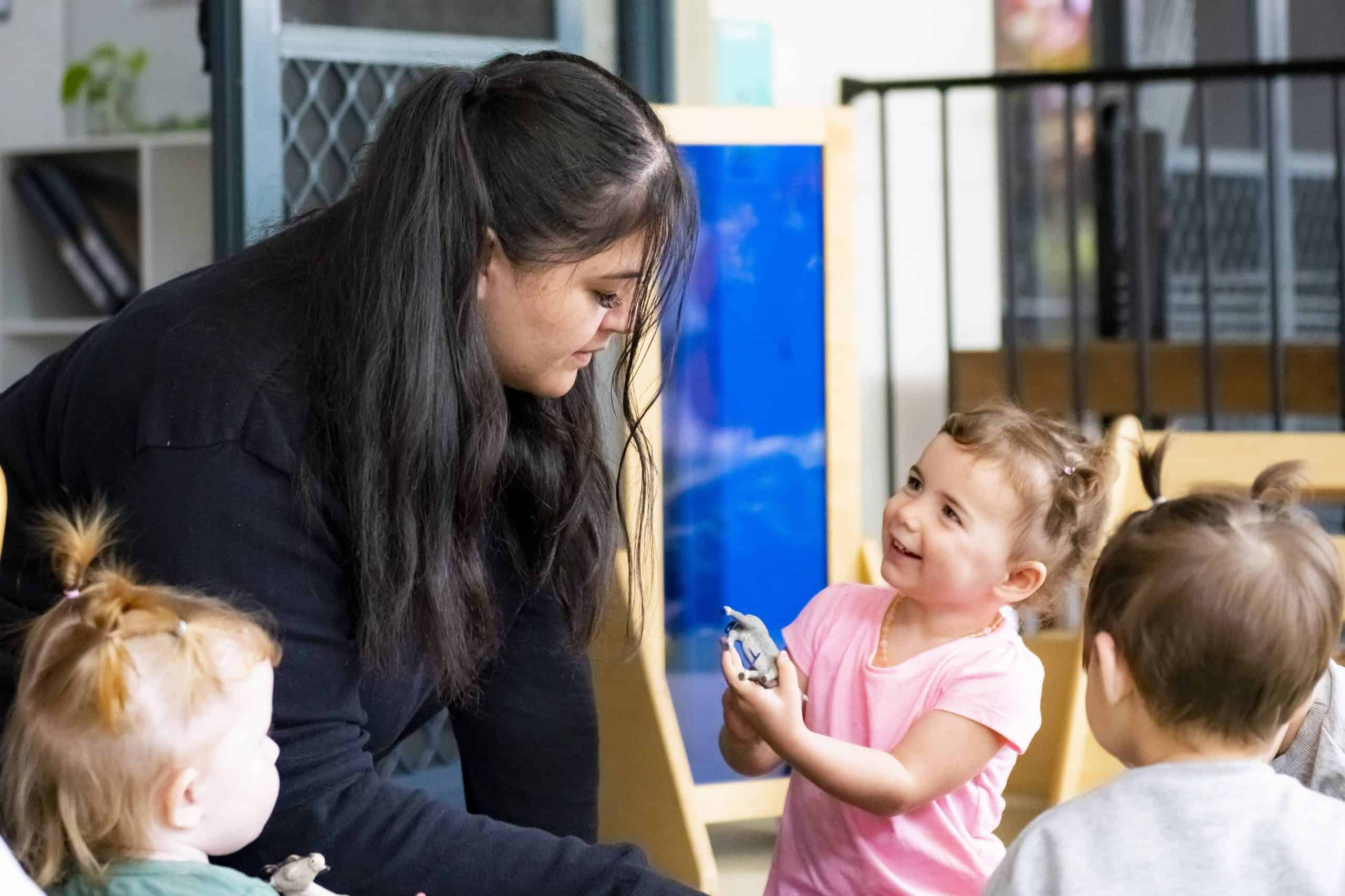 An adult woman interacts with three young children in a daycare room. Two are seated while the standing child, dressed in pink, smiles and holds a toy. The other two children observe attentively.