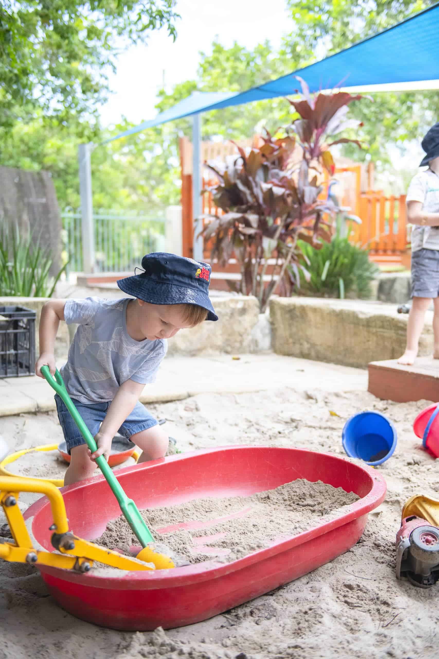 A young child wearing a hat is playing with a yellow and green toy digger in a sandpit, filling a red tray with sand, at an outdoor kindergarten playground. Other toys and children, engaged in early learning activities, are visible in the background.