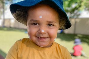 A young child wearing a blue hat and yellow shirt, with sunscreen on their face, smiles at the camera while playing outside at daycare. Another child is seen in the blurred background sitting on the grass.