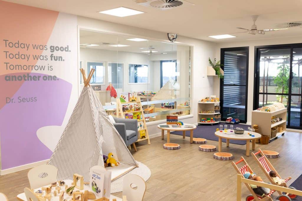 A children's playroom designed for early learning, featuring various toys, books, and seating areas. A teepee tent is set up in the room. A wall displays a Dr. Seuss quote, adding charm to the space. Large windows and bright lighting ensure a welcoming environment perfect for day care activities.