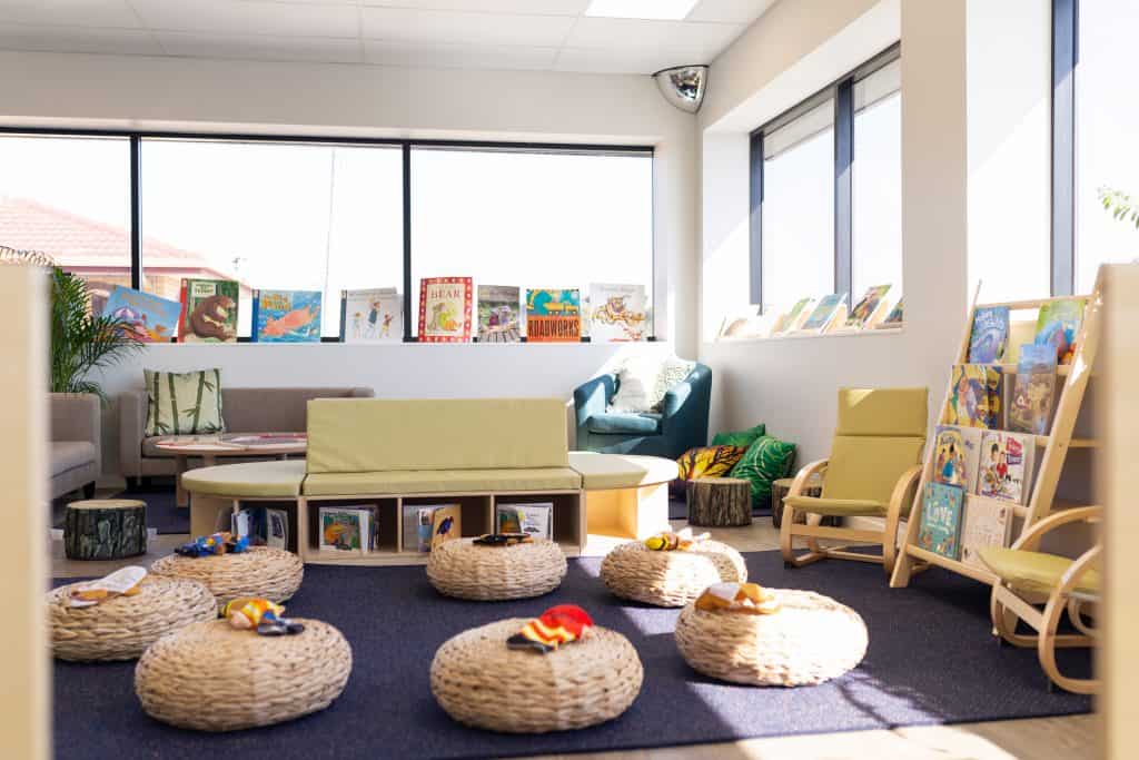 A brightly lit, cozy children's reading area with low beige seating, round straw cushions, and books displayed on wall-mounted shelves and nearby stands. Perfect for a day care or kindergarten setting, large windows allow natural light to fill the space.