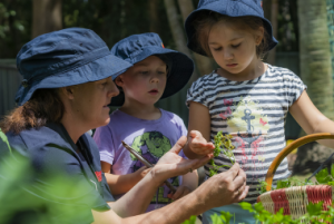 An adult shows two children plants in a garden while all three wear navy blue hats. The adult, involved in early learning, holds a plant in their hand, and the children observe closely.