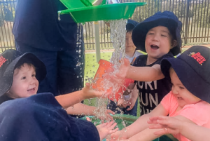 Four young children wearing hats play with water at a playground, joyfully catching the flow from a green water trough. This playful and interactive outdoor activity highlights early learning experiences often seen in kindergarten and childcare settings.